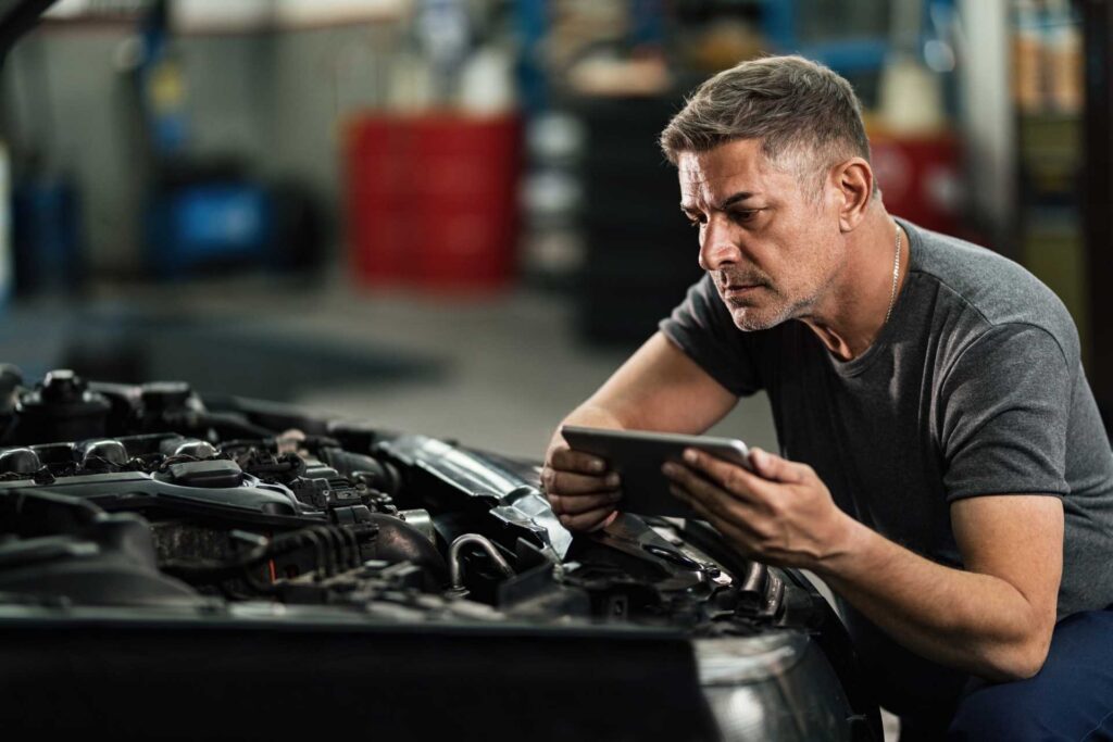 garage problems, male mechanic holding tablet crouched beside car